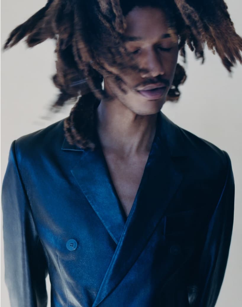 A person with thick, textured dreadlocks moves their head slightly, eyes closed. They wear a dark blue double-breasted blazer against a light background